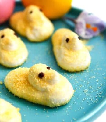 Yellow vegan peeps on a blue plate with colored Easter eggs behind it.