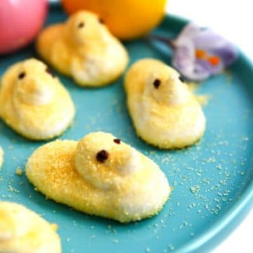 Yellow vegan peeps on a blue plate with colored Easter eggs behind it.