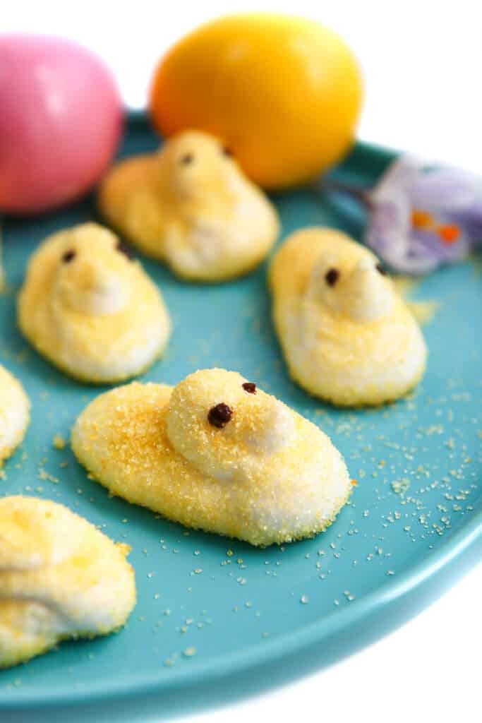 Homemade vegan marshmallow peeps on a blue plate with a flower and colored Easter eggs.