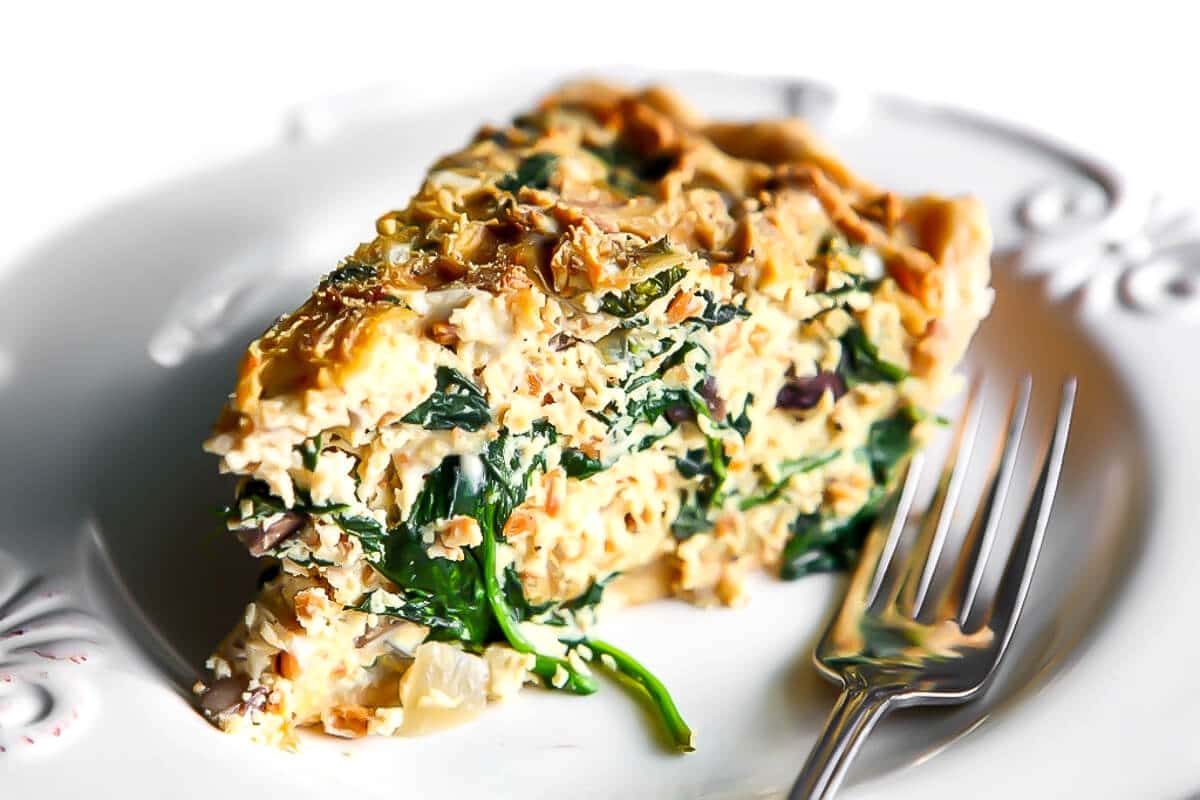A slice of quiche with spinach, mushrooms, bacon bits and vegan cheese in it on a white plate.