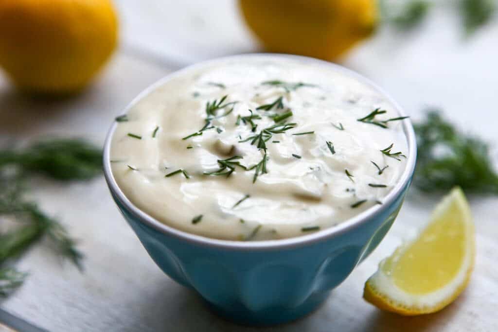 A small blue bowl filled with homemade vegan tartar sauce with dill and lemon on the side.