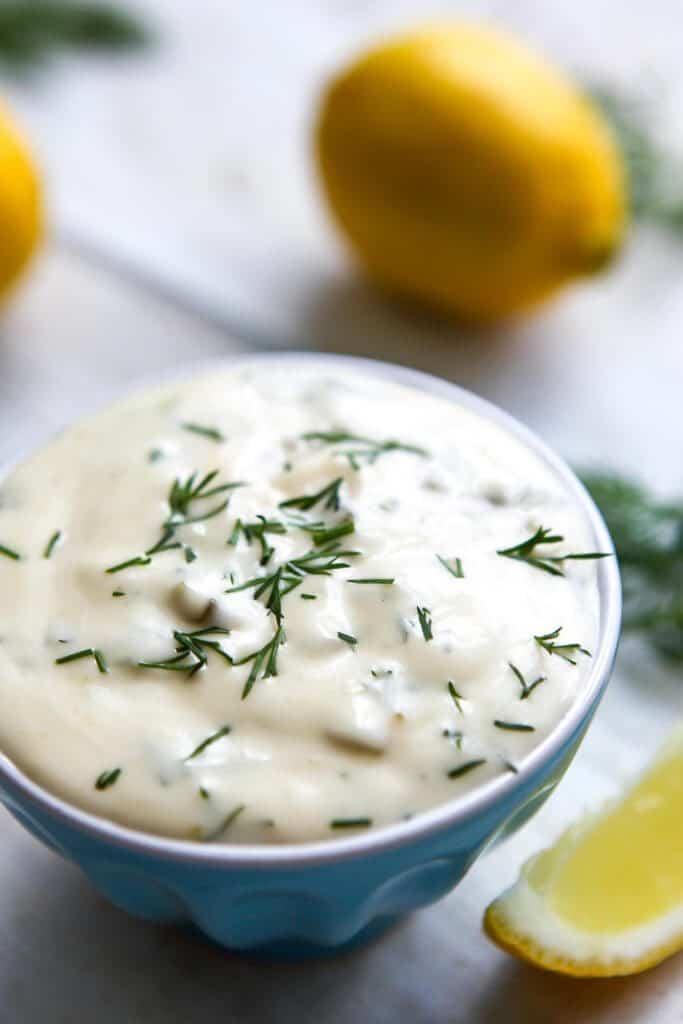 A blue bowl filled with homemade vegan tartar sauce with lemons and dill around it. 