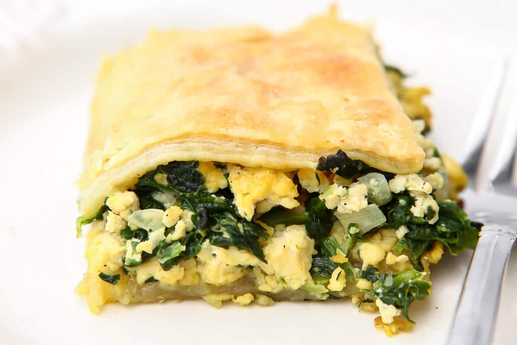 A piece of Spanakopita with tofu and spinach in it on a white plate with a fork on the side.