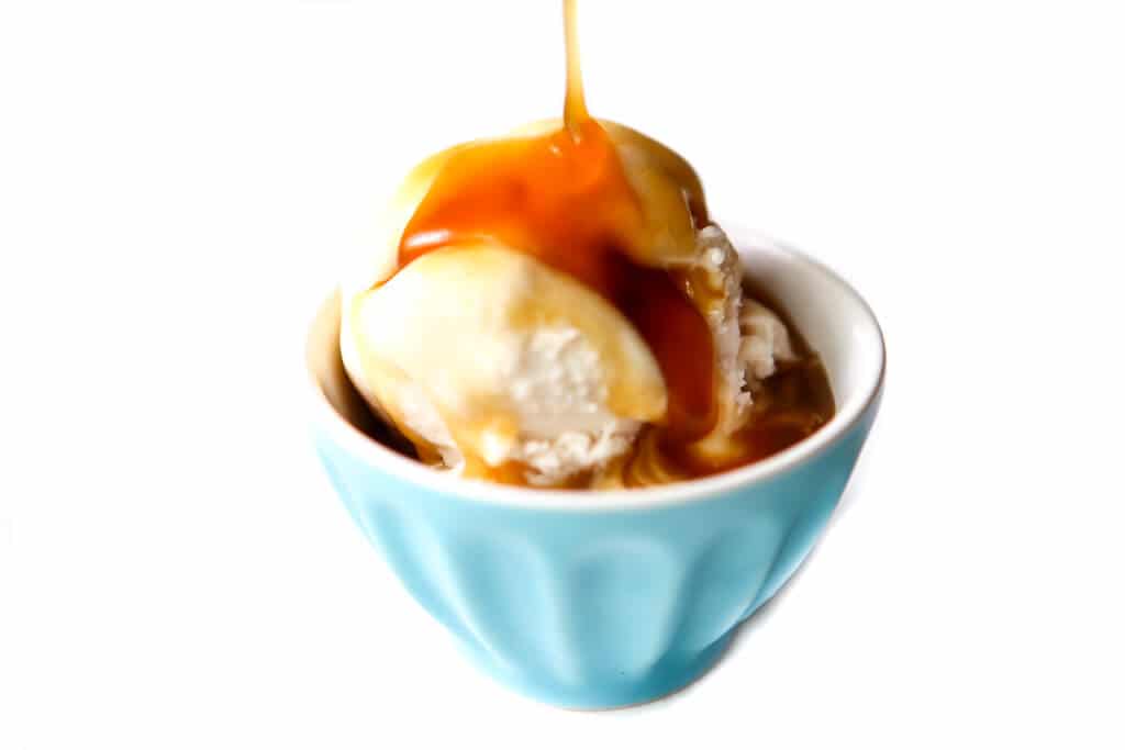 A blue bowl filled with ice cream with vegan butterscotch sauce being poured over top.