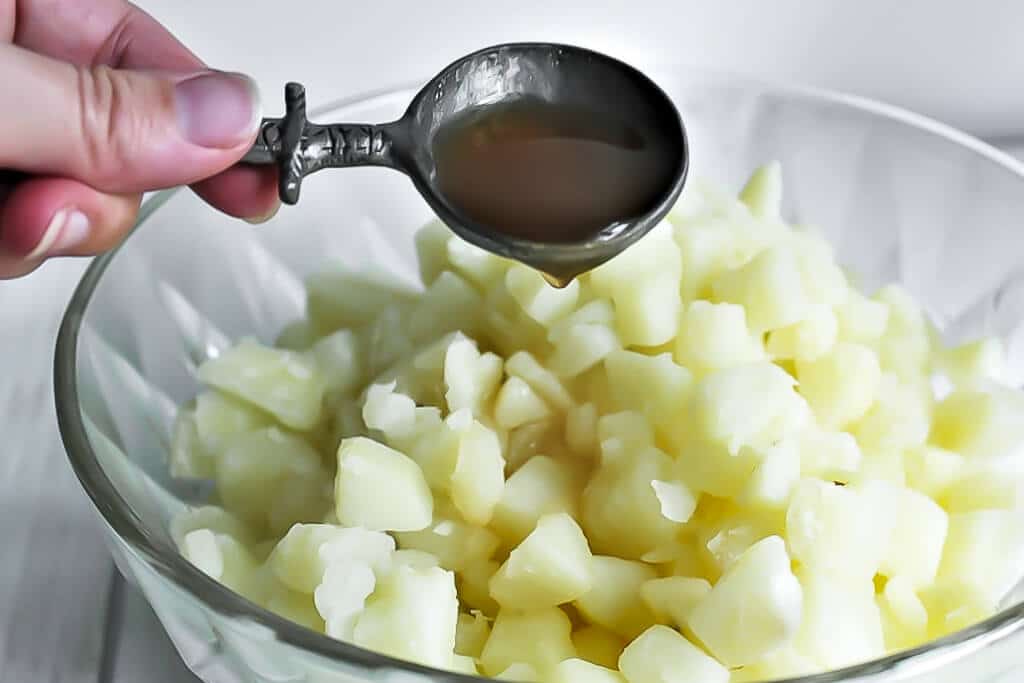 A spoon with apple cider vinegar is held above above a bowl of cooked, chopped potatoes.