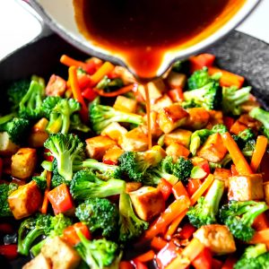 Teriyaki tofu with vegetables in a frying pan with teriyaki sauce being poured over it.