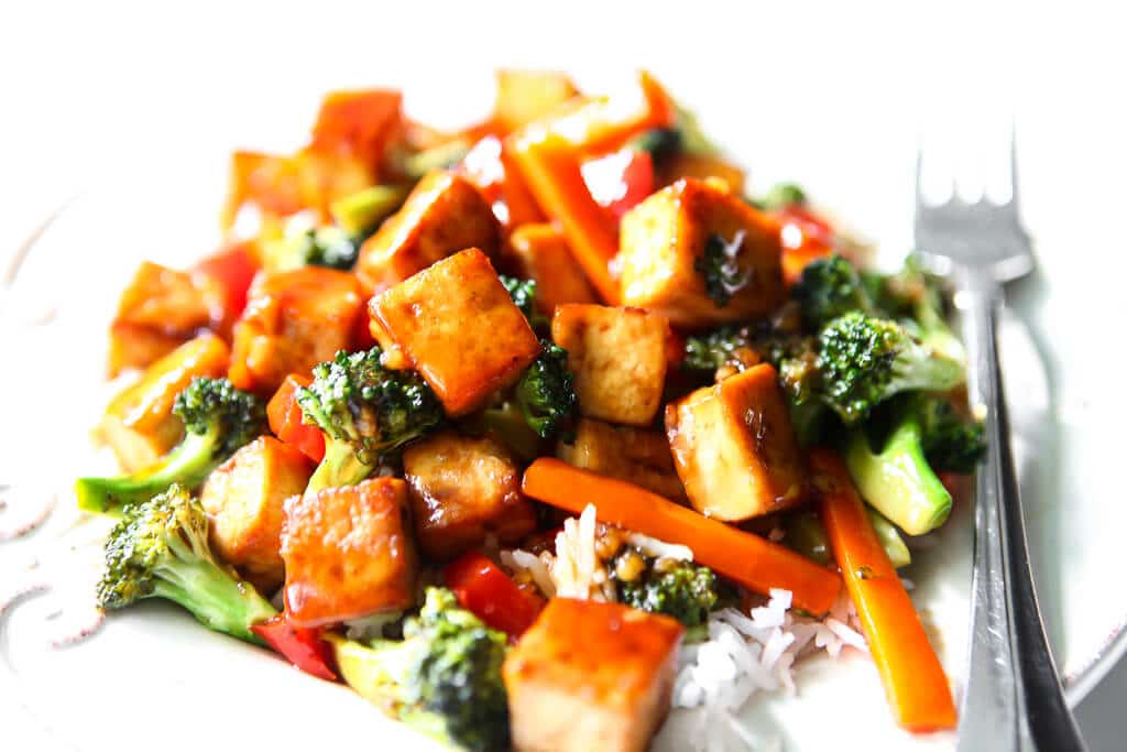 A white plate filled with vegan teriyaki tofu and stir-fried veggies served over rice. 