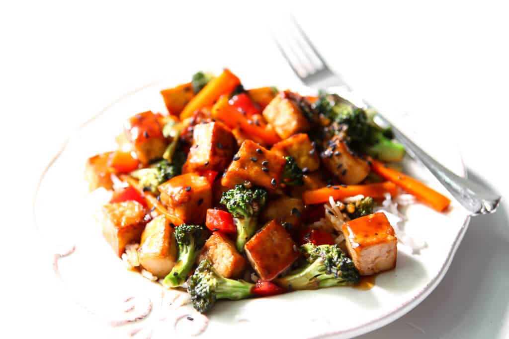 A white plate with teriyaki tofu stir fry served over rice and topped with sesame seeds. 