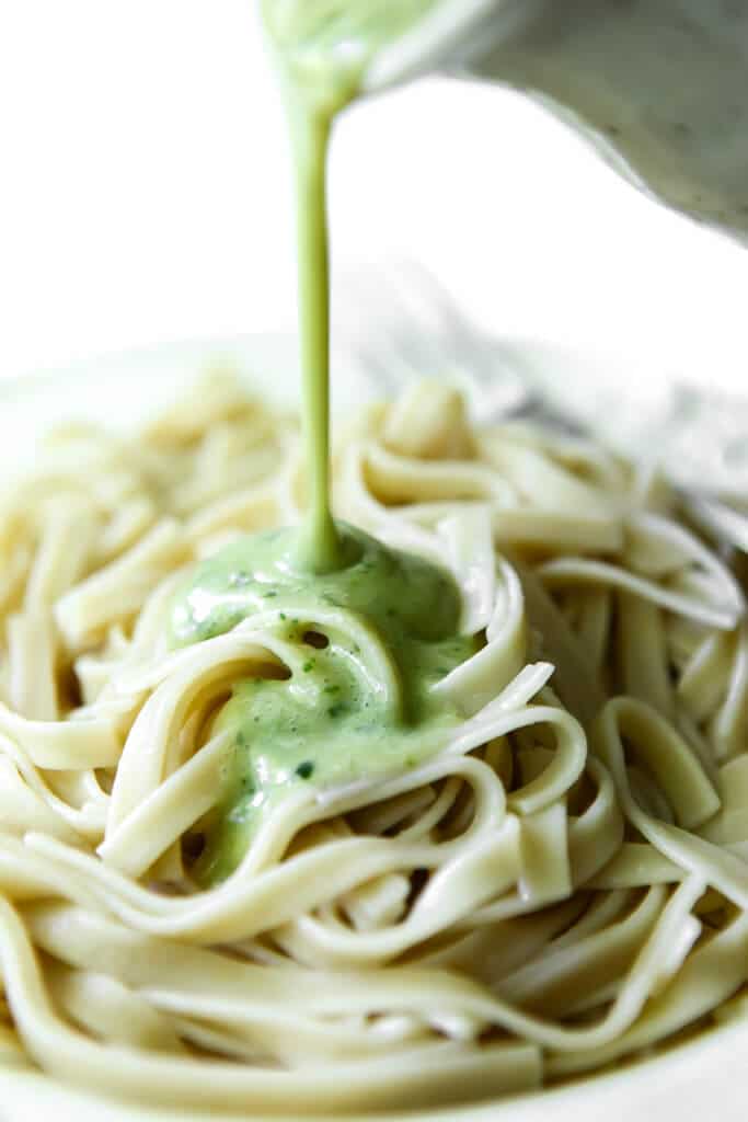 Creamy pesto pasta sauce being poured over a plate of gluten-free linguine pasta.