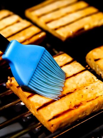 Four slices of grilled tofu on a barbecue grill being brushed with a marinade.