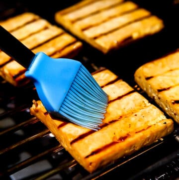 Four slices of grilled tofu on a barbecue grill being brushed with a marinade.