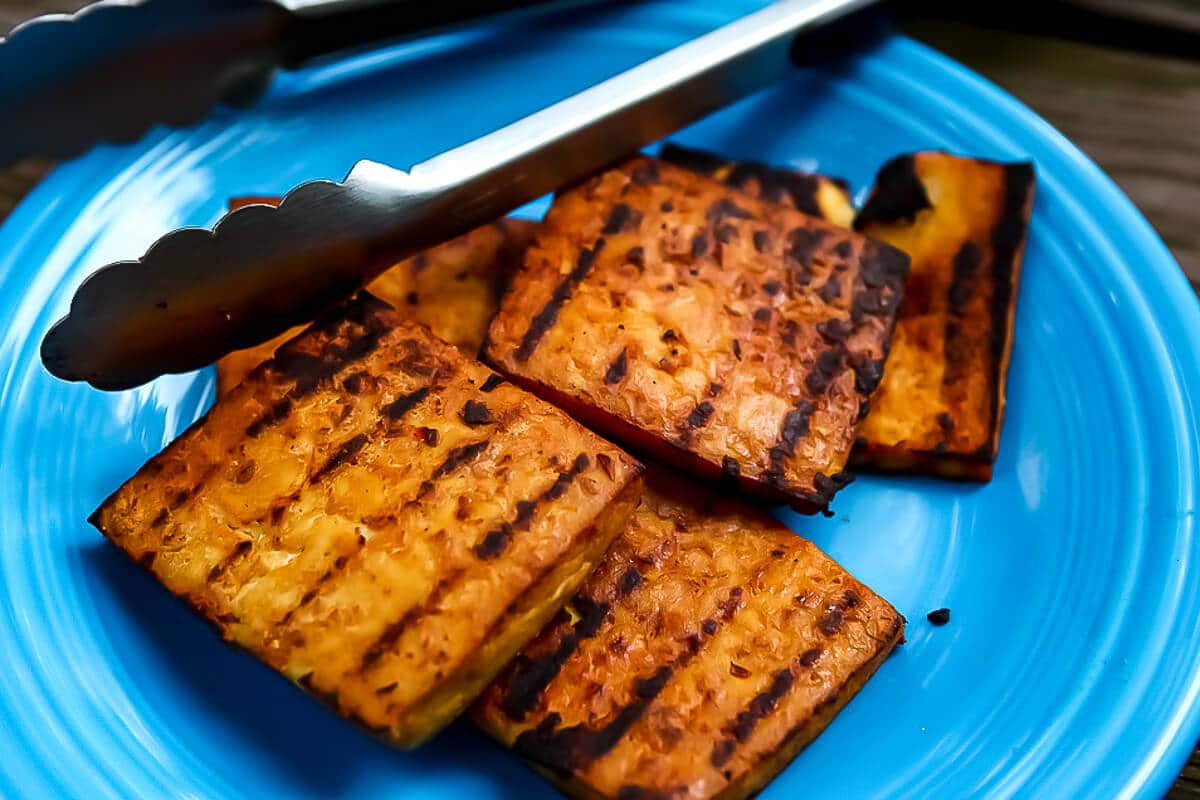 Marinated grilled tofu on a bright blue plate with tongs on the side.