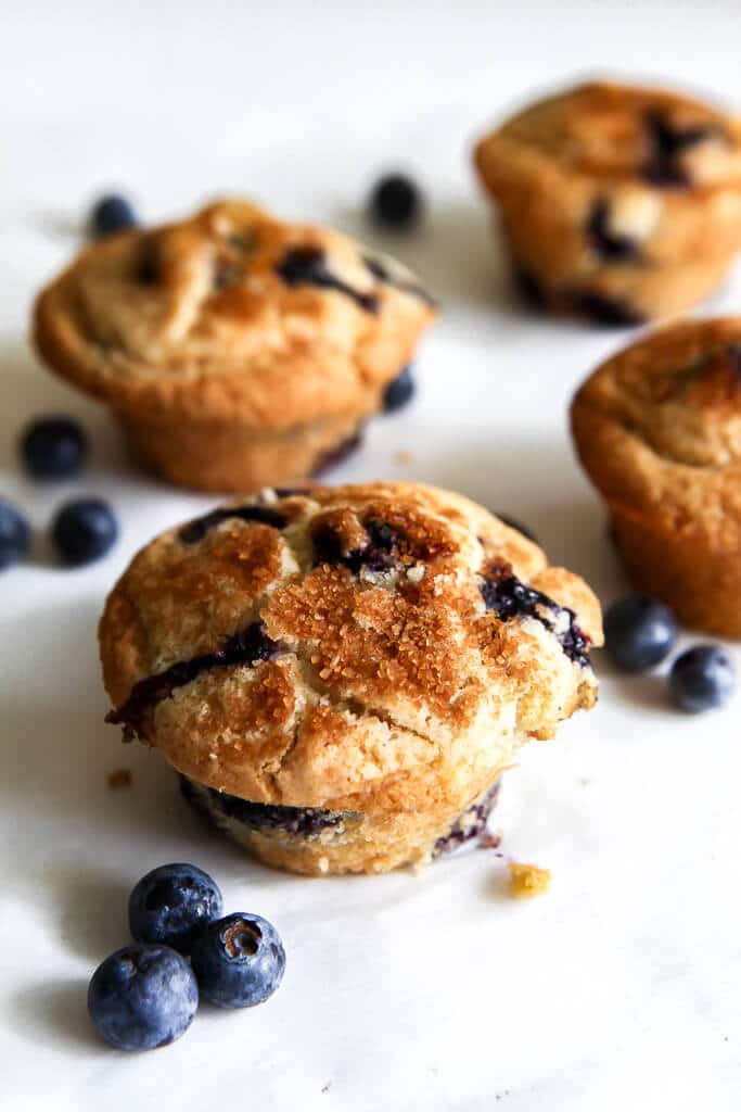 Four vegan blueberry muffins on a white background with fresh blueberries around it.