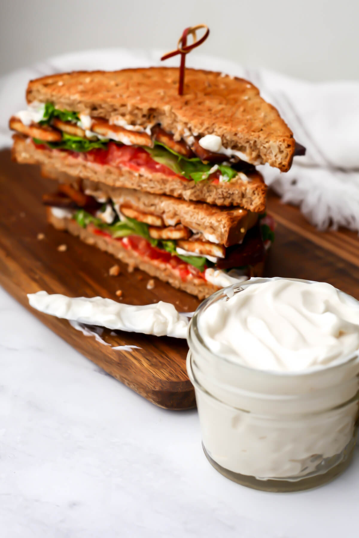 A jar of vegan mayo in front of a sandwich with a knife on the side.