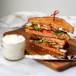 A jar of vegan mayo next to a vegan sandwich with a knife to spread the mayo.