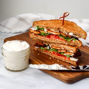 A jar of vegan mayo next to a vegan sandwich with a knife to spread the mayo.