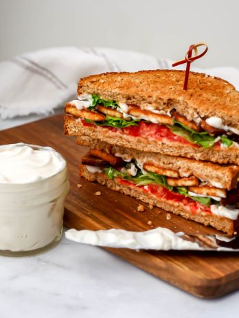 A jar of vegan mayo next to a vegan sandwich with a knife to spread the mayo.
