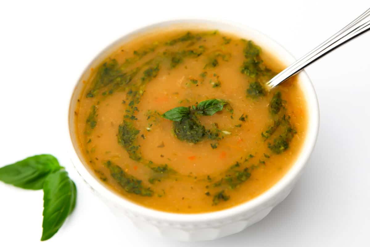 A bowl of creamy vegan soup with a swirl of pesto on top and a basil leaf on the side.