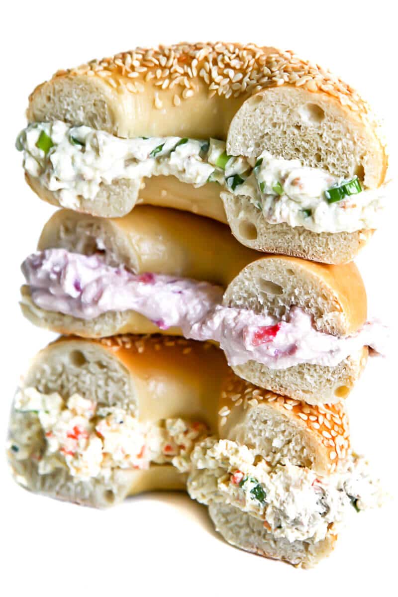A stack of 3 bagels with herb, strawberry, and garden veggie style tofu cream cheese in the middle of each bagel.