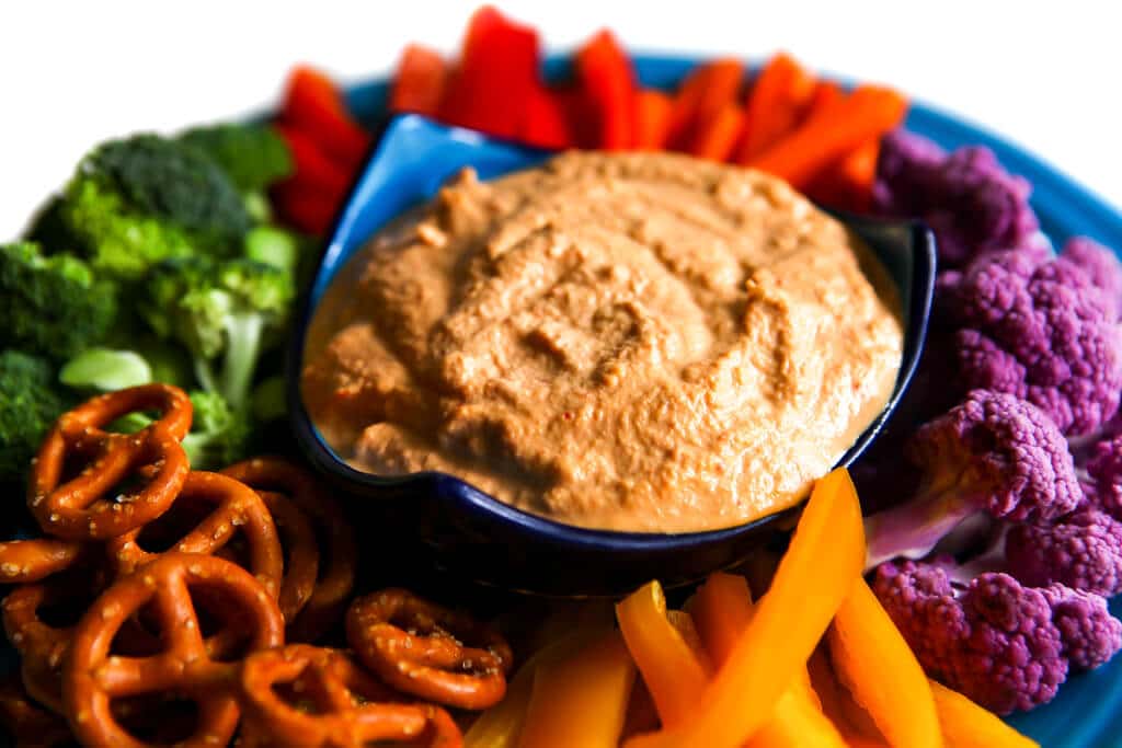 A colorful assortment of veggies on a blue plate with a bowl of red pepper cashew dip in the center.