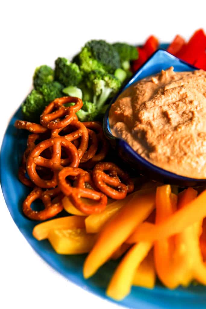An assortment of veggies and pretzels on a blue plate with vegan cashew cheese dip in the middle.