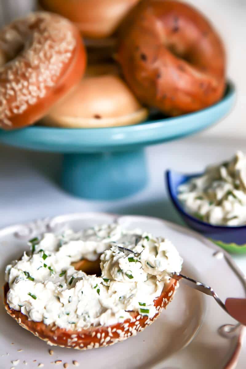 A bagel with vegan garlic and chives cream cheese with a blue plate filled with bagels behind it.