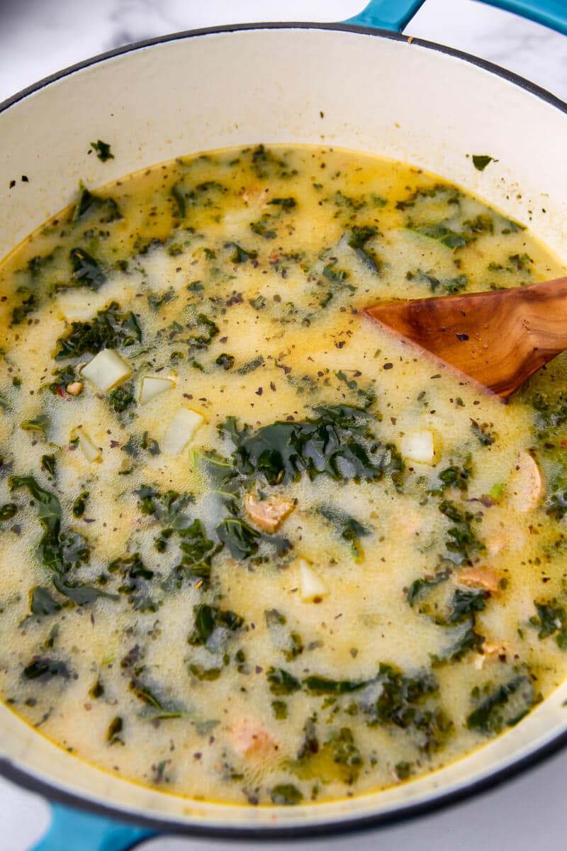 A blue dutch oven filled with vegan zuppa toscana being stirred with a wooden spoon.