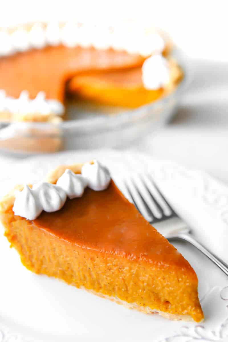 A slice of vegan pumpkin pie on a white plate with a fork next to it with a full pie behind it.