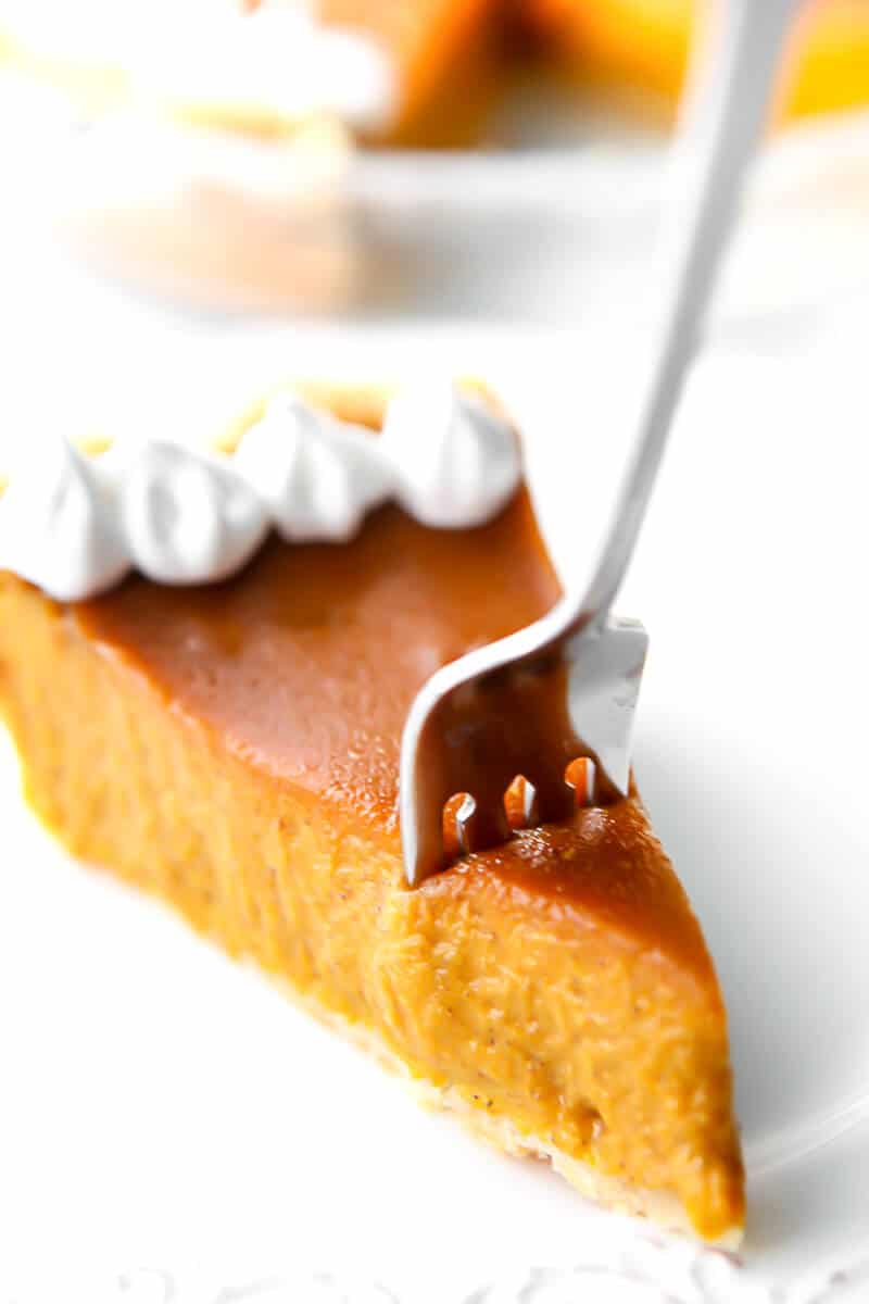 A slice of pumpkin pie with a fork in it with a pie dish behind it.