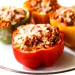 Four stuffed peppers on a plate filled with quinoa, sauce and vegan meat, topped with vegan cheese.