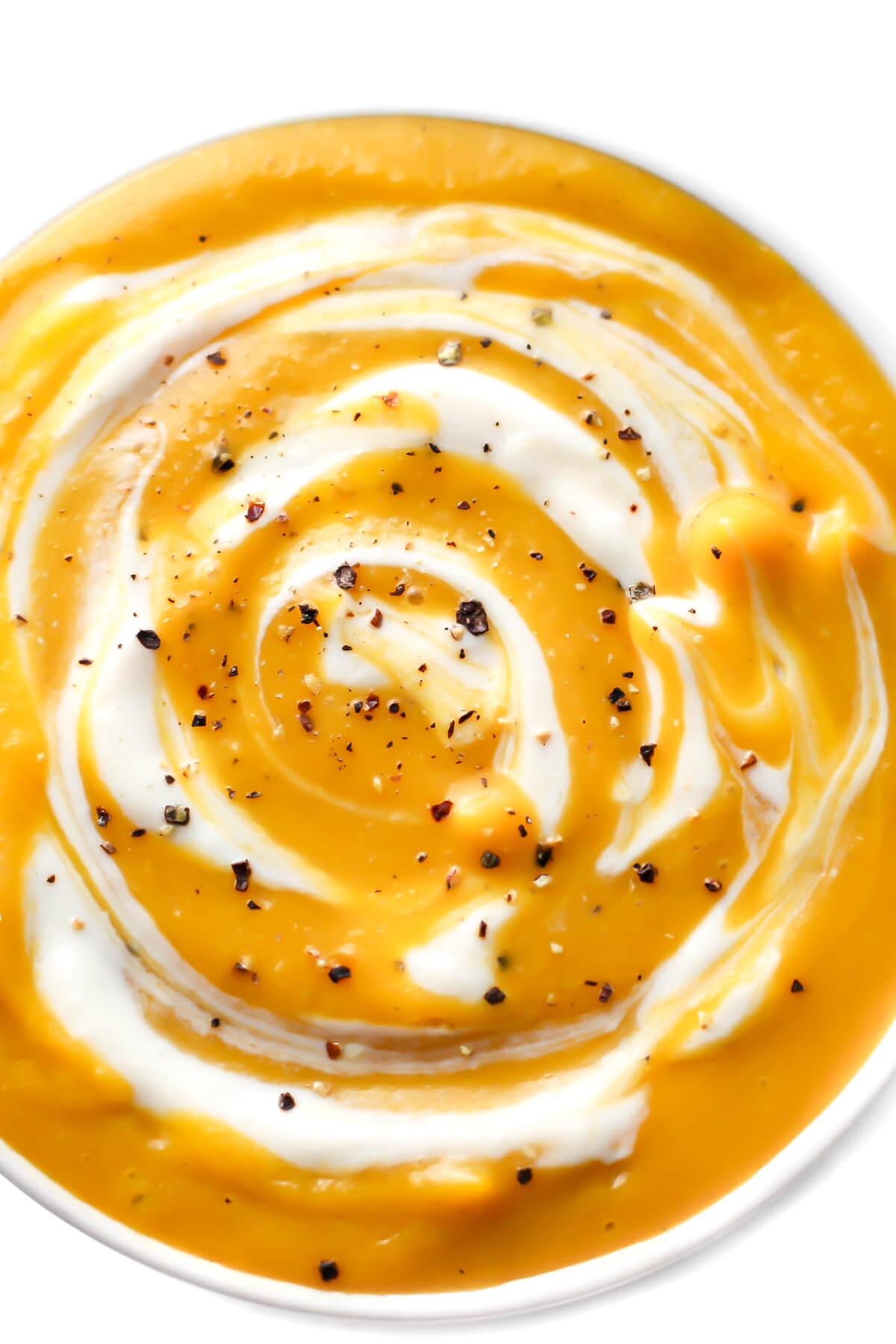 A close up of a white bowl filled with creamy vegan butternut squash soup with some vegan sour cream and ground pepper swirled on top.