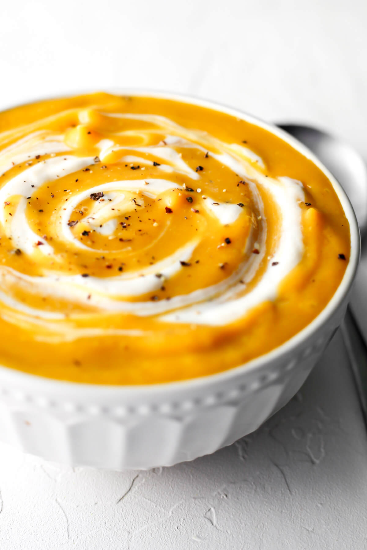 A white bowl filled with creamy vegan butternut squash soup with sour cream swirled on top and a spoon on the side.