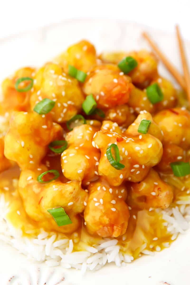 A top view of a white plate with crispy orange cauliflower with sesame seeds and green onions sprinkled on top 