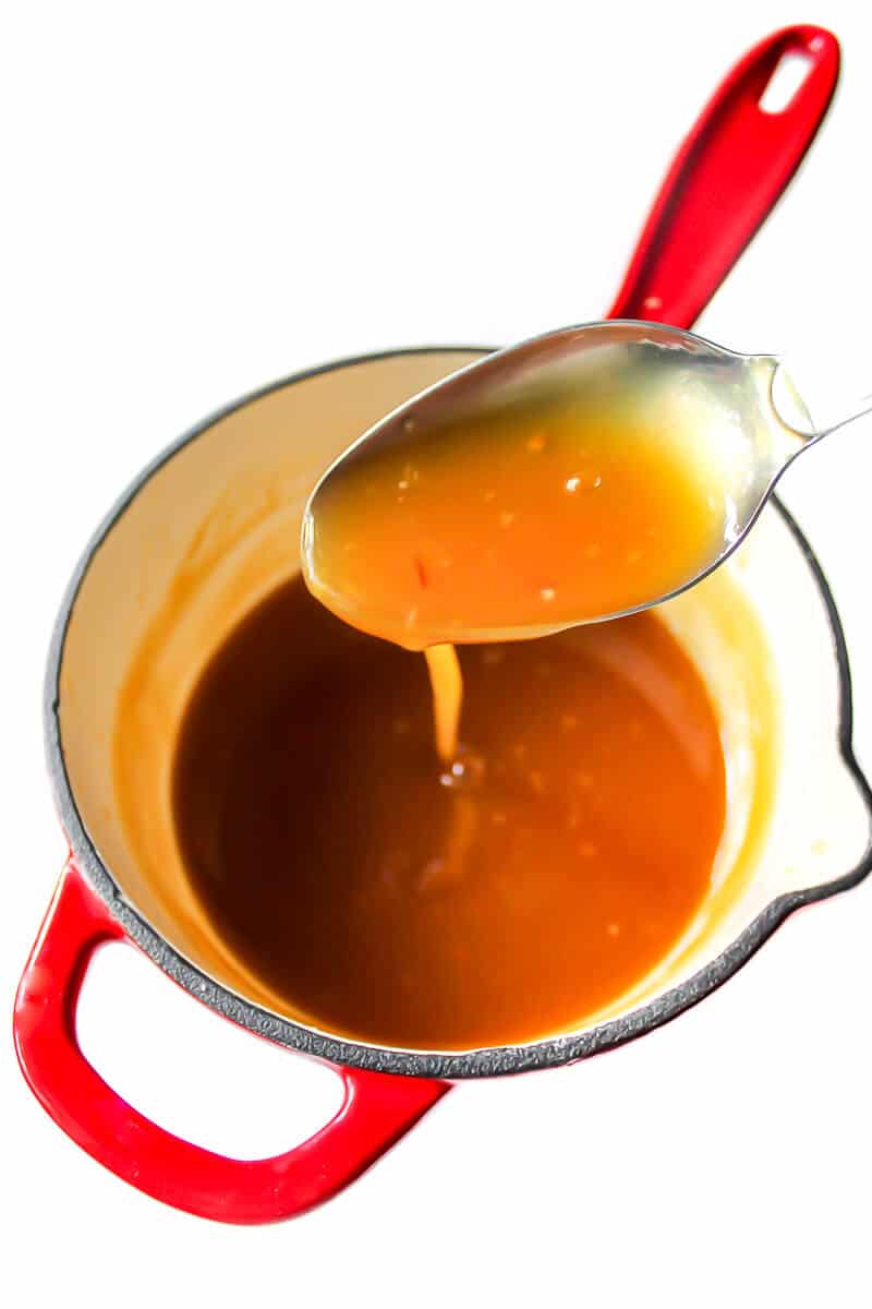 A dripping spoon full of vegan and gluten free orange sauce over a red saucepan.