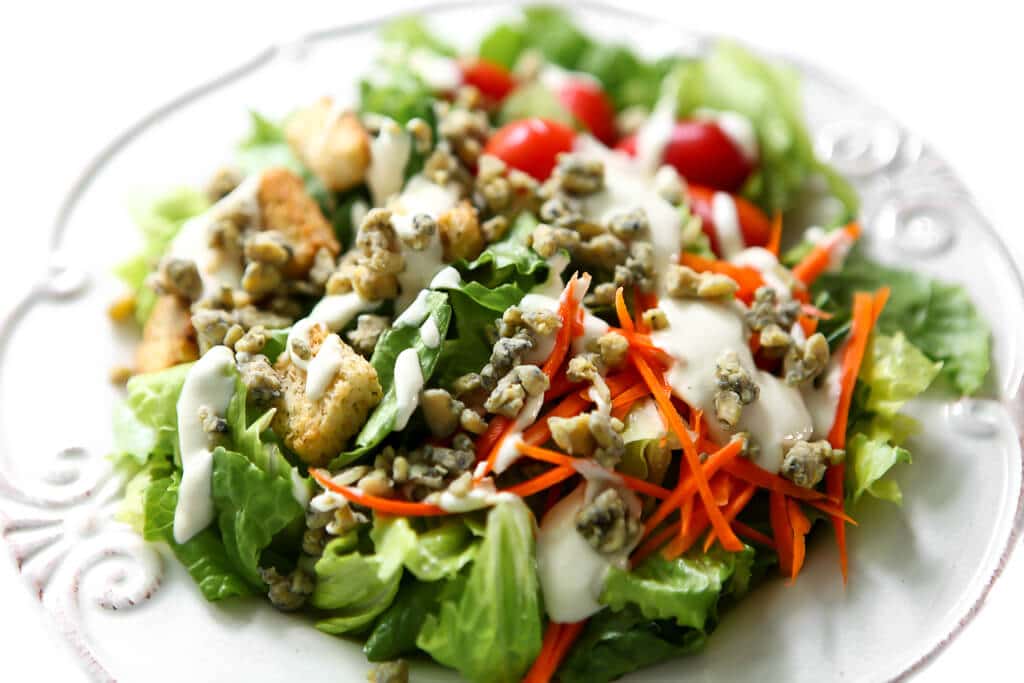 A white plate full of salad with vegan blue cheese creamy dressing and blue cheese crumbles on top.