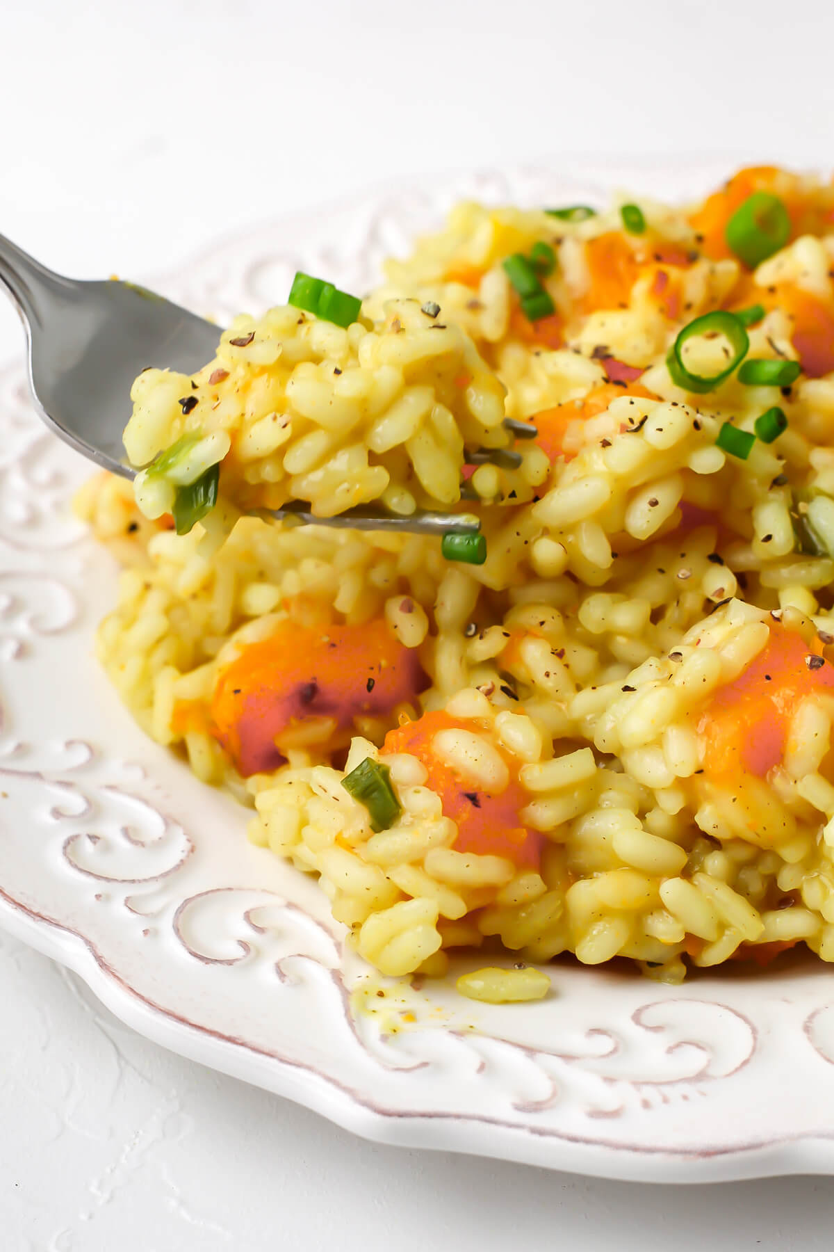 A white plate full of creamy vegan butternut squash risotto topped with slices of green onion and black pepper with a fork in it.