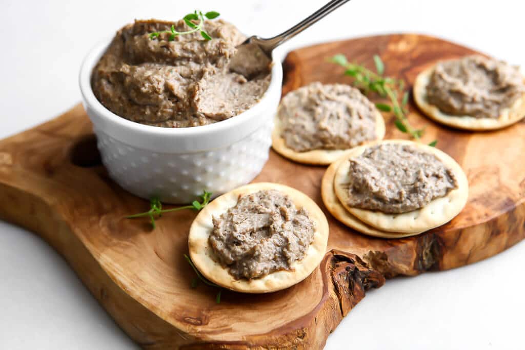 A small bowl of mushroom pate on a cutting board with crackers topped with vegan pate around it. 