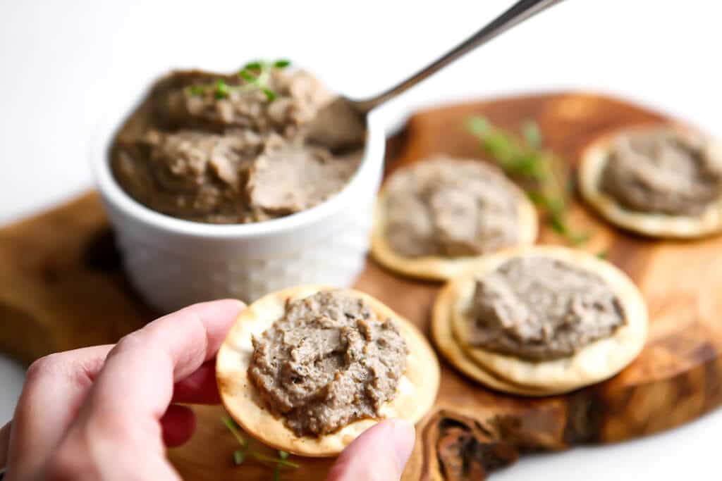 Someone holding a cracker with pate spread on it with more cracker with vegan pate behind it. 