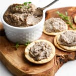 A small bowl of vegan mushroom pate on a cutting board with crackers topped with vegan pate around it.