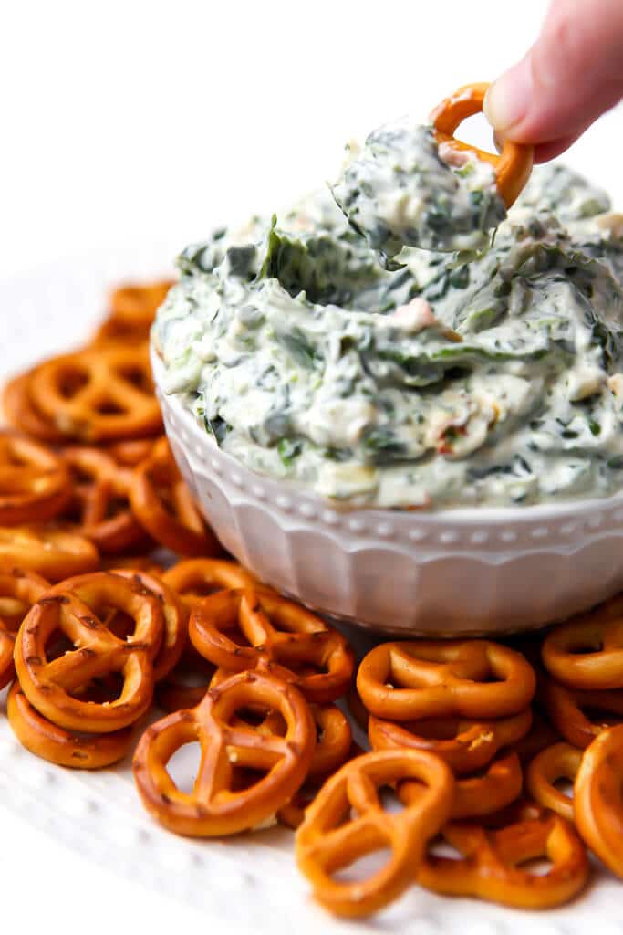 Someone dipping a pretzel into a bowl of vegan spinach dip.