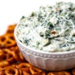A close up of vegan spinach dip in a white bowl with pretzels around it.