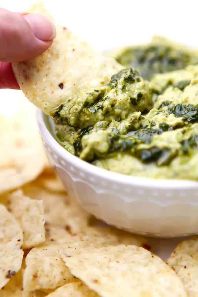 A bowl of vegan pesto hummus in a white bowl with someone dipping a chip into it. 