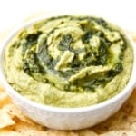 A white bowl filled with pesto hummus with extra pesto swirled on top.