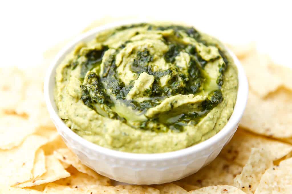 A white bowl filled with pesto hummus with chips around it.