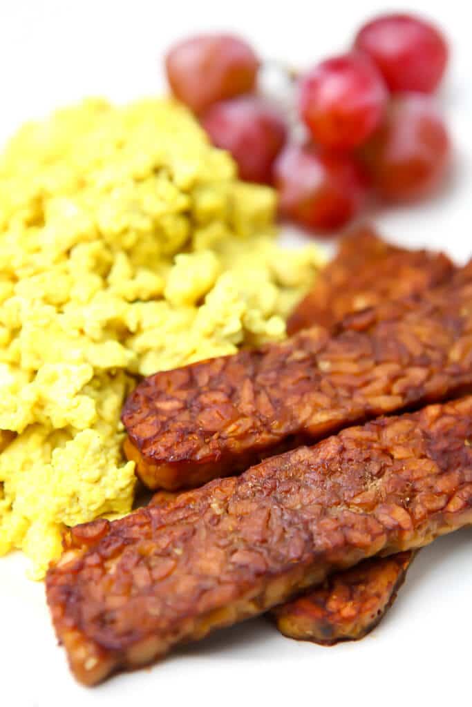 Vegan bacon made from tempeh served on a plate with vegan scrambled eggs and red grapes.