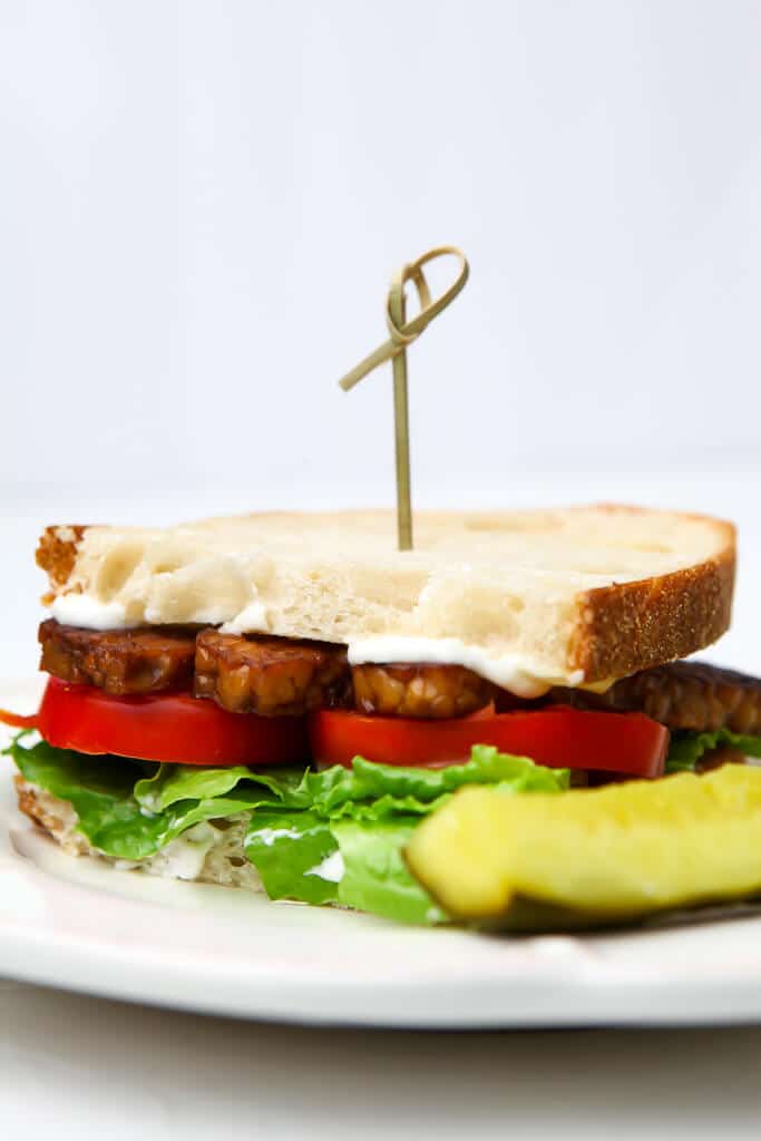 A vegan BLT sandwich made with tempeh bacon on a white plate with a pickle on the side.
