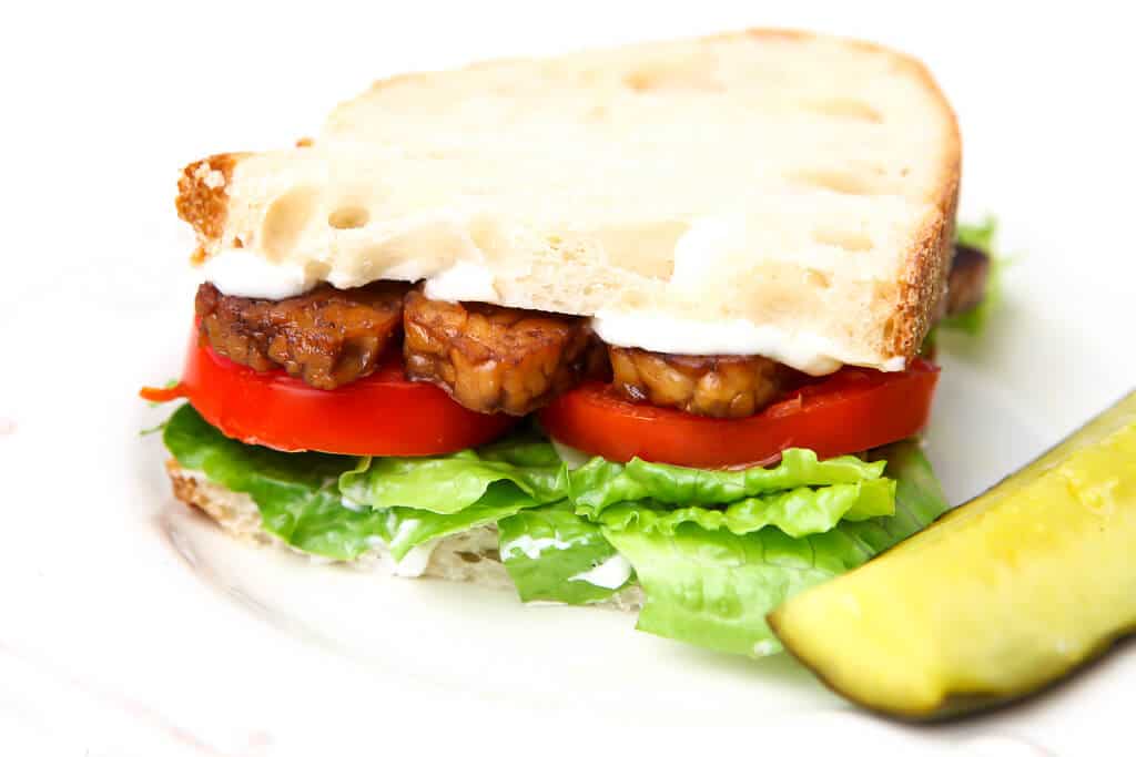 A vegan BLT sandwich made with tempeh bacon with a pickle on the side. 