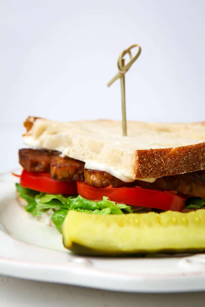 A vegan bacon, lettuce and tomato sandwich on a white plate with a pickle on the side. 