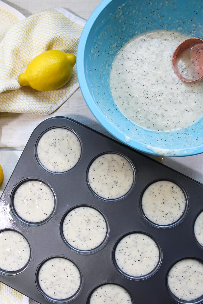 A muffin tin filled with the batter for lemon poppy seed muffins with a blue mixing bowl next to it.