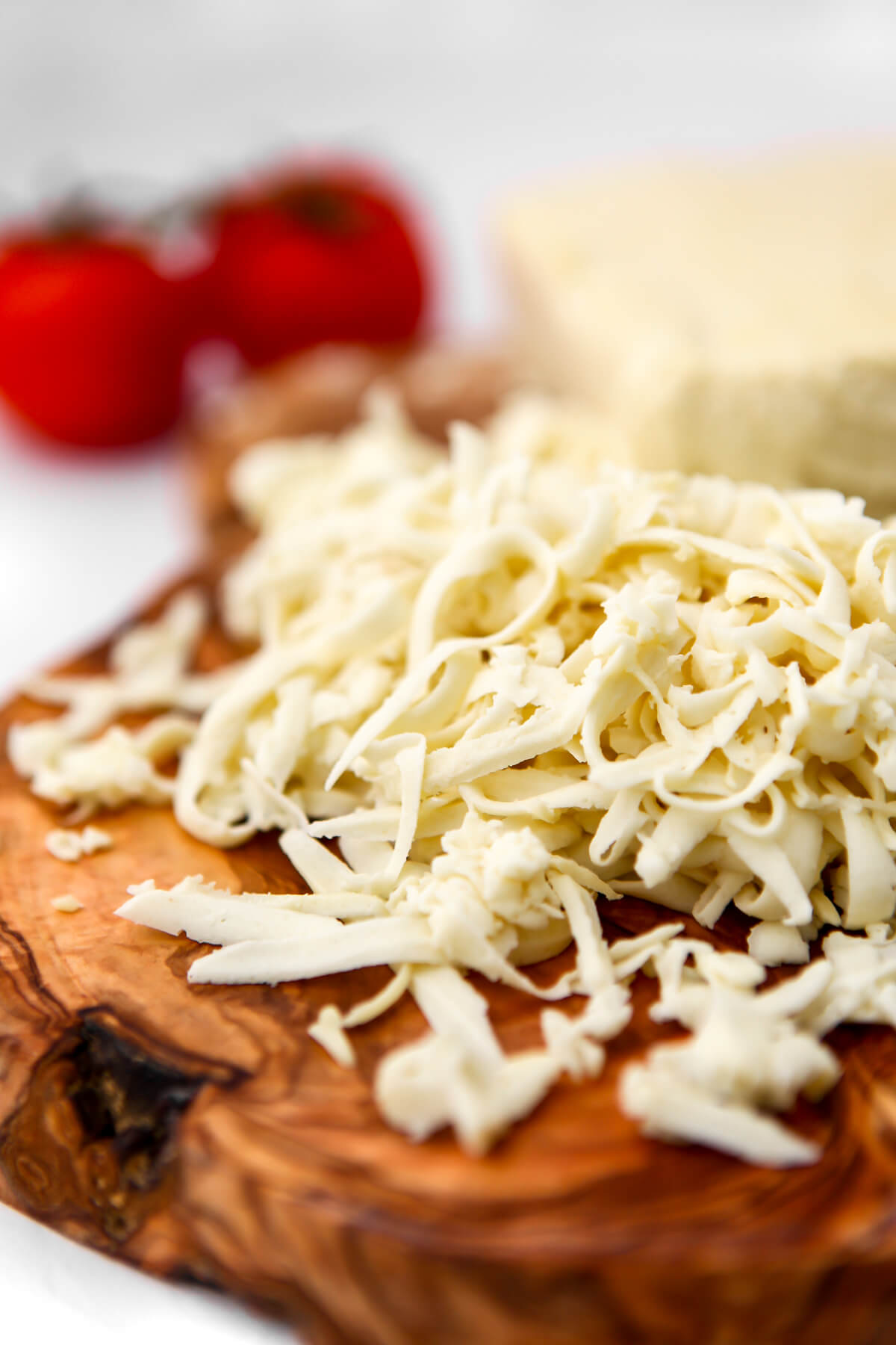 Shredded vegan mozzarella cheese on a wooden cutting board with two tomatoes behind it.